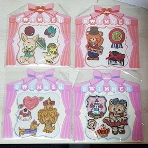 Puede incluir: Cuatro tarjetas rosas y blancas con la palabra "SWIMMER" impresa en ellas. Cada tarjeta presenta un conjunto diferente de parches termoadhesivos con varios diseños, incluyendo un conejito, un oso, un perro y un castillo. Los parches son coloridos y presentan detalles bordados.