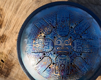May include: A blue steel handpan with a decorative pattern and multiple indentations. The handpan is resting on a wooden surface.