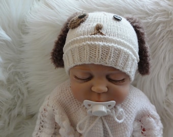 May include: A white knitted baby hat with brown fur ear flaps and a brown felt dog face design. The hat is on a baby's head.