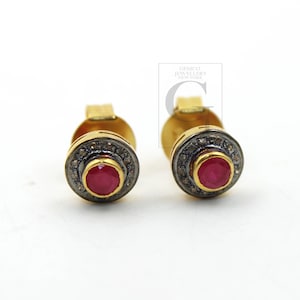 May include: A pair of gold stud earrings with a red ruby gemstone in the center of each earring. The ruby is surrounded by a halo of small diamonds.