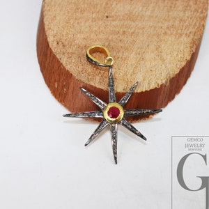 Peut inclure: Un pendentif en argent en forme d'étoile avec des diamants noirs et un rubis rouge au centre. Le pendentif a une bélière en or et est sur une surface en bois.