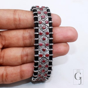 Può includere: Un bracciale in argento con una fascia nera intrecciata, ornato da pietre preziose rosse e piccoli diamanti bianchi. Il bracciale presenta un design geometrico con file alternate di pietre rosse e dettagli di diamanti.