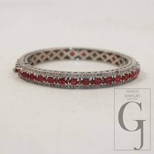 May include: A hinged bangle bracelet featuring a row of oval-cut red stones set in a dark metal frame. The band is accented with small pavé-set stones along the edges and a geometric lattice pattern on the sides. Includes a GEMCO JEWELRY NEWYORK logo.