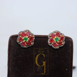 May include: A pair of gold earrings with a flower design. The earrings are set with red and green gemstones and black diamonds.