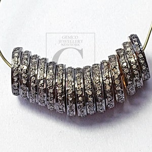 May include: A collection of dark silver ring beads, each set with tiny, glittering stones. The beads are threaded onto a thin, gold-coloured wire, forming a curved line against a white backdrop. These beads are likely for jewellery making.