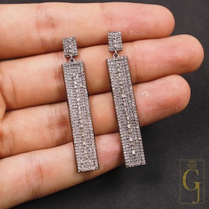 May include: A pair of silver-toned rectangular drop earrings. Each earring features a small square top and a long, rectangular body, both encrusted with small, sparkling stones. The earrings are displayed against a neutral background.
