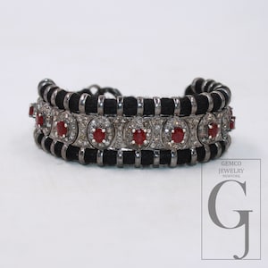 May include: A black and silver bracelet with red gemstones. The bracelet features a woven black band with silver accents and circular settings with red stones and surrounding clear stones. The bracelet is displayed on a white surface.