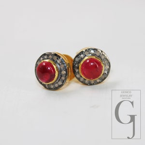 Peut inclure: Une paire de boucles d'oreilles à clous rondes avec une pierre précieuse rouge centrale taillée en cabochon, entourée d'un halo de petits diamants pavés sertis sur fond sombre. Les montures sont en or avec une finition métallique oxydée. Texte : GEMCO JEWELRY NEWYORK G.