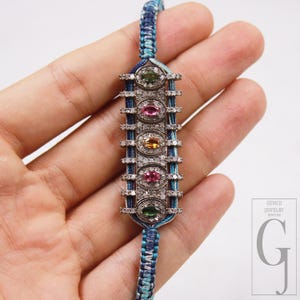 May include: A silver bracelet with a series of oval-shaped gemstones in green, pink, and yellow, interspersed with small clear stones. The bracelet is attached to a blue and teal braided cord. The Gemco Jewelry logo is in the lower right corner.