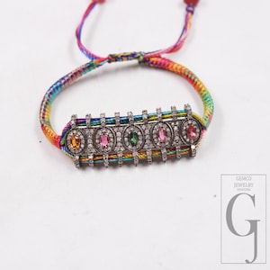 May include: A colorful bracelet with a rainbow-colored woven band. The bracelet features a silver-toned rectangular centerpiece with five oval-shaped gemstones in yellow, pink, green, and pink. The centerpiece is encrusted with small, clear stones.
