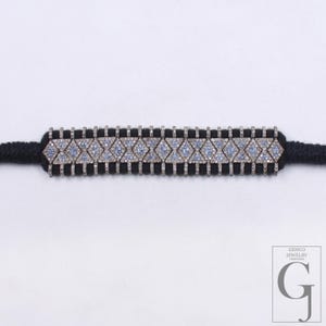 Puede incluir: Una pulsera plateada con un diseño geométrico con piedras preciosas azules y transparentes. La pulsera está engarzada en un cordón negro. El logotipo de Gemco Jewelry New York está en la esquina inferior derecha.