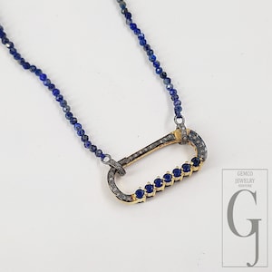 Può includere: Una collana con un grande pendente ovale con pietre blu e piccole pietre scintillanti. Il pendente è incorniciato in metallo color oro. La collana ha una catena di perline di piccole perline blu scuro.