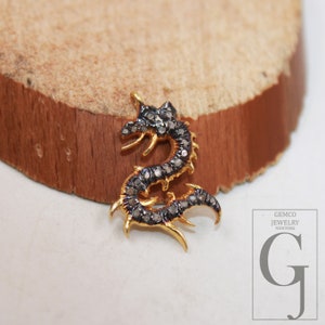 Può includere: Un ciondolo a forma di drago in oro con diamanti neri. Il ciondolo è su una superficie di legno. GEMCO JEWELRY NEW YORK è stampato su uno sfondo bianco.