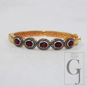 May include: Gold bangle bracelet with five oval-shaped red gemstones surrounded by smaller clear gemstones.
