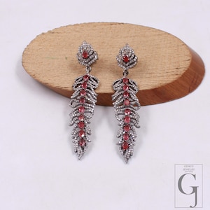 May include: A pair of silver-toned dangle earrings with a leaf-like design. Each earring features a cluster of red gemstones and is accented with small, clear stones. The earrings are displayed on a wooden surface.