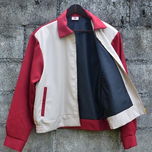 May include: A red, cream, and navy blue zip-up jacket hanging on a wooden hanger. The jacket has red sleeves and collar, cream-colored front panels, and a navy blue interior lining. A small red pocket is visible on the left side.
