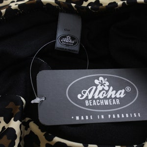 K&ouml;nnte beinhalten: Ein schwarz-wei&szlig;er Leoparden-Badeanzug mit einem schwarzen Etikett, auf dem "Aloha Beachwear" und "Made in Paradise" steht.