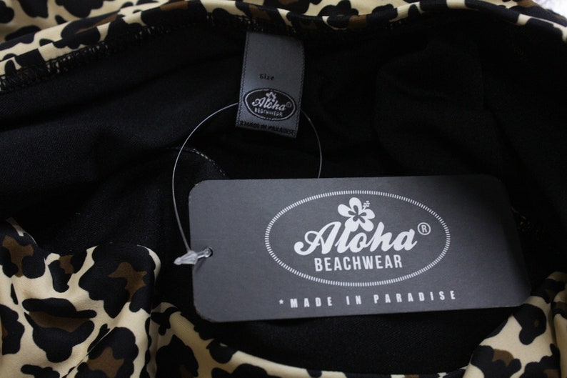 K&ouml;nnte beinhalten: Ein schwarz-wei&szlig;er Leoparden-Badeanzug mit einem schwarzen Etikett, auf dem "Aloha Beachwear" und "Made in Paradise" steht.