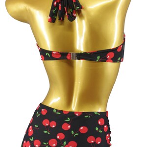 May include: Black and red cherry print bikini with a halter top and high-waisted bottoms. The bikini is on a gold mannequin. The brand name "Aloha Beachwear" is printed on the bottom of the bikini.
