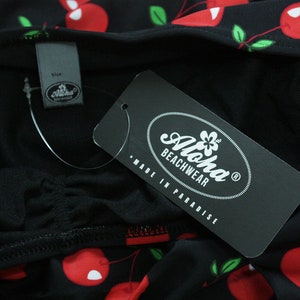 May include: Black and white tag with the text "Aloha Beachwear" and "Made in Paradise". The tag is attached to a black fabric with a red cherry print.