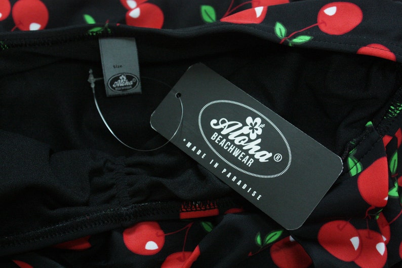 May include: Black and white tag with the text "Aloha Beachwear" and "Made in Paradise". The tag is attached to a black fabric with a red cherry print.
