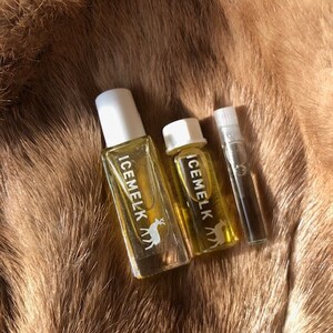May include: Three glass bottles of perfume with white labels and a deer logo. The labels read "ICEMELK". The bottles are on a brown fur background.