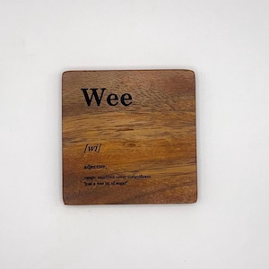 May include: A square wooden coaster with a natural brown grain pattern. The text printed in black reads: Wee, [wi], adjective, usage: small but never insignificant, "just a wee bit of sugar". The coaster has rounded corners and a smooth finish.