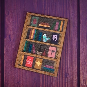 Puede incluir: Una estantería de madera con cinco estantes llenos de libros, algunos artículos decorativos y un pequeño objeto azul de cristal. La estantería está pintada de color marrón claro y tiene un borde marrón oscuro.