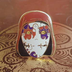 May include: A gold-coloured enamel pin featuring a stylised face with a red, white and purple mask. The mask has a stitched design and two large eyes with yellow and purple irises.
