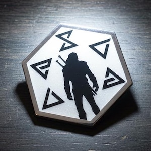 May include: A hexagonal white and gray sticker featuring a black silhouette of a figure with a sword on their back. The sticker has black geometric symbols around the figure. The sticker is on a dark wooden surface.