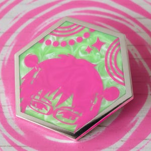 Puede incluir: Un espejo compacto hexagonal plateado con un diseño de personaje de anime rosa sobre un fondo verde. El espejo compacto está sobre un diseño de remolino rosa contra un fondo de vetas de madera blanca.