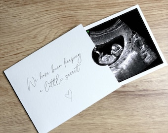 May include: A white card with the text "We have been keeping a little secret" and a small heart symbol. An ultrasound image of a fetus is partially visible. The card is on a wooden surface.