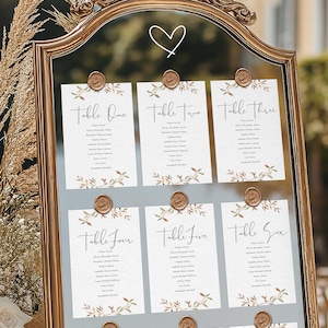 Peut inclure: Un miroir doré affiche six plans de table de mariage. Chaque carton blanc présente une calligraphie élégante, des numéros de table et des noms d'invités. Des éléments floraux et des sceaux de cire ajoutent une touche de sophistication.