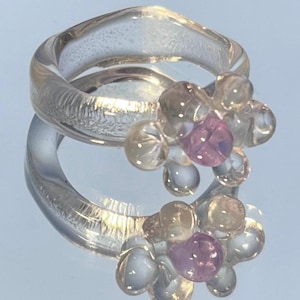 Könnte beinhalten: Zwei transparente Harzringe mit floralem Design. Jeder Ring hat eine Ansammlung klarer und rosa Harzperlen. Die Ringe spiegeln sich auf einer glänzenden Oberfläche und erzeugen einen Spiegeleffekt. Die Ringe haben ein einzigartiges, künstlerisches Design.