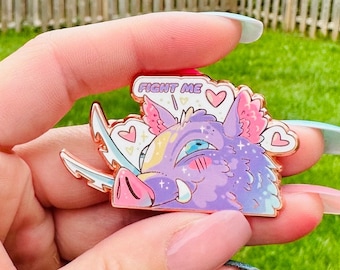 May include: A purple and pink enamel pin of a cartoon boar with a pink snout and a single blue eye. The boar has a speech bubble above its head that says "Fight Me!" with a pink heart next to it.