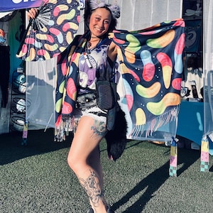 May include: A person wearing a black and white kimono with a colorful gummy worm pattern. The kimono has a black fur trim and is worn over a black tank top. The person is holding a black and white fan with a colorful gummy worm pattern. The person is standing in front of a white tent with a sign that says "Creative Paradise".