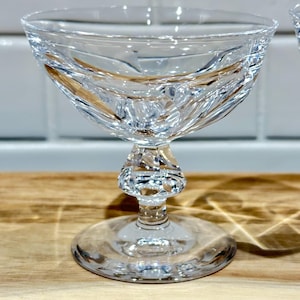 May include: A clear, cut-glass champagne coupe with a wide bowl and a short stem. The bowl features a swirling, textured design. The coupe sits on a round base, and is displayed on a wooden surface.