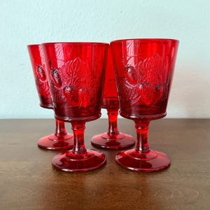 May include: A set of four ruby red glass goblets featuring a raised, embossed pattern of strawberries and leaves on the exterior. Each goblet has a short, sturdy stem and a wide, circular base, crafted from translucent, vibrant red pressed glass.