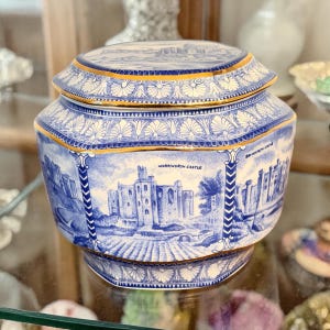May include: A blue and white ceramic jar with a lid, featuring a detailed illustration of Warkworth Castle. The jar has a gold trim and a decorative pattern around the edges. The overall design is vintage and ornate.