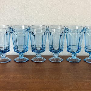 May include: Six vintage blue water goblets with a faceted design. The glasses have a hexagonal base and a stem. The goblets are arranged in a row on a wooden surface.