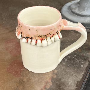 Może przedstawiać: Ceramiczny kubek o unikalnym designie. Górna część kubka ma różowy pasek z rzędem białych zębów. Kubek ma biały korpus i zakrzywiony uchwyt. Kubek stoi na odblaskowej powierzchni.