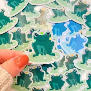 May include: A pile of frog stickers in shades of green and blue. Each sticker depicts a cartoon frog with a crown, sitting on a lily pad. One sticker is held up by a hand. The stickers are suitable for crafts.