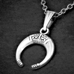 May include: A silver-toned crescent-shaped pendant suspended from a matching link chain. The pendant features a smooth, curved design with a textured, engraved band across the top section and small, etched markings at both pointed ends.