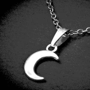 May include: A silver-toned crescent moon pendant suspended from a matching silver-toned metal link chain. The pendant features a smooth, polished surface that reflects light, and it is attached to the chain by a simple, tapered metal bail.