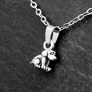 May include: A silver-toned metal necklace featuring a small, three-dimensional pendant shaped like a sitting puppy. The pendant is attached to a delicate link chain, showcasing a detailed canine figure with floppy ears and a rounded body design.