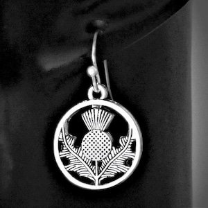 May include: A silver-toned metal earring featuring a circular frame that encloses a detailed thistle design. The earring hangs from a simple hook-style wire, showcasing a textured, oxidised finish that highlights the intricate floral pattern.