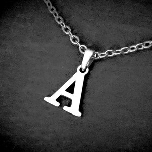 May include: A silver-coloured necklace featuring a pendant shaped like the letter "A". The pendant is suspended from a chain. The background is a dark, textured surface. The necklace is in sharp focus.