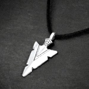 May include: Silver-colored arrowhead pendant necklace on a black cord. The arrowhead pendant has a detailed design and is suspended from a black cord. The background is a dark, textured surface.