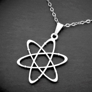 May include: Silver-coloured pendant necklace with an atom symbol design. The pendant depicts a stylized atom structure, with intersecting orbital rings and small bead-like details. The necklace has a delicate chain.