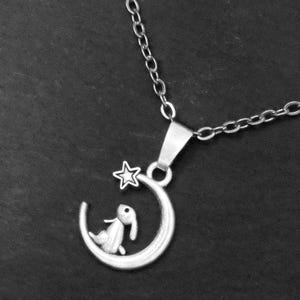 May include: A silver-toned metal necklace featuring a pendant shaped like a crescent moon. A small rabbit figure sits inside the curve of the moon, and a five-pointed star is attached to the upper tip of the crescent. The pendant hangs from a chain.